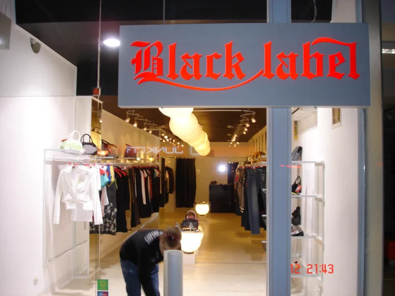 Вход на магазин за дрехи с надпис Black Label и витрина, показваща подредени закачалки с облекла и интериорно осветление.