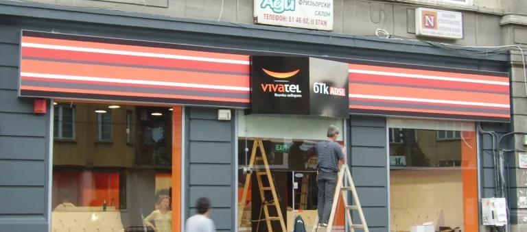 Работник на стълба монтира брандирана табела с логото на Vivatel и БТК ADSL над входа на търговски обект.