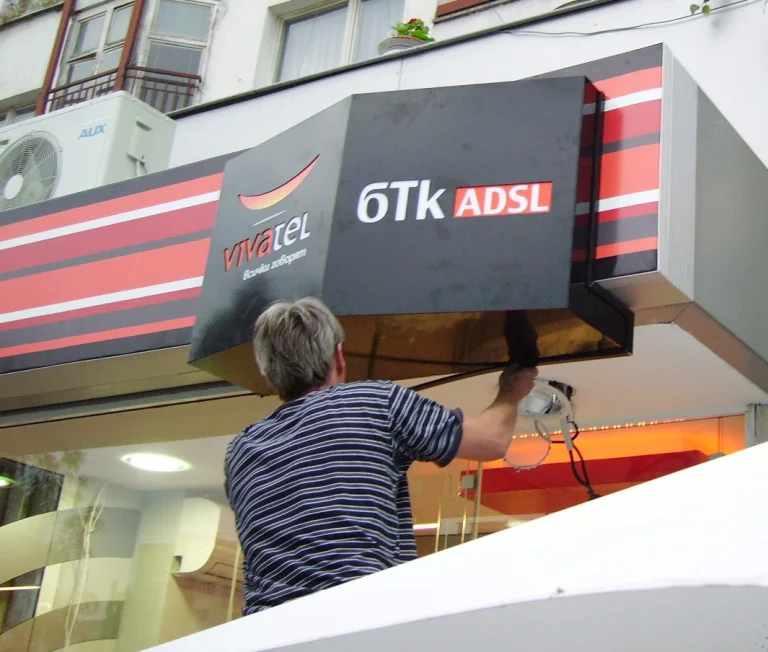 Техник монтира светеща рекламна табела с логото на Vivatel и надпис 6Tk ADSL над входа на търговски обект.