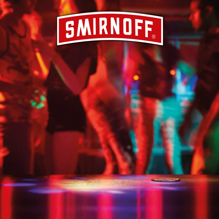Логото на Smirnoff е поставено върху размазан фон от нощен клуб с танцуващи хора и цветно осветление.