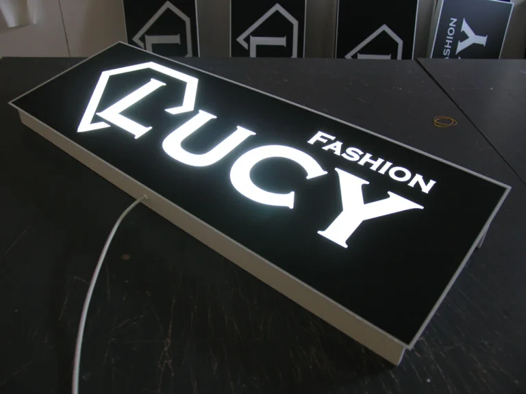 Светеща правоъгълна табела с надпис LUCY FASHION и графичен елемент, поставена върху работна маса.
