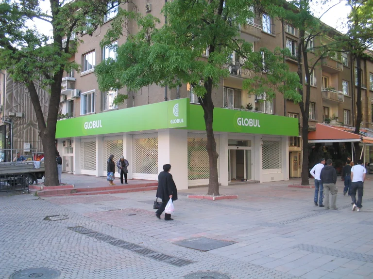 Ъглов магазин с яркозелена брандирана табела с логото на GLOBUL, разположен на градска улица с пешеходци.