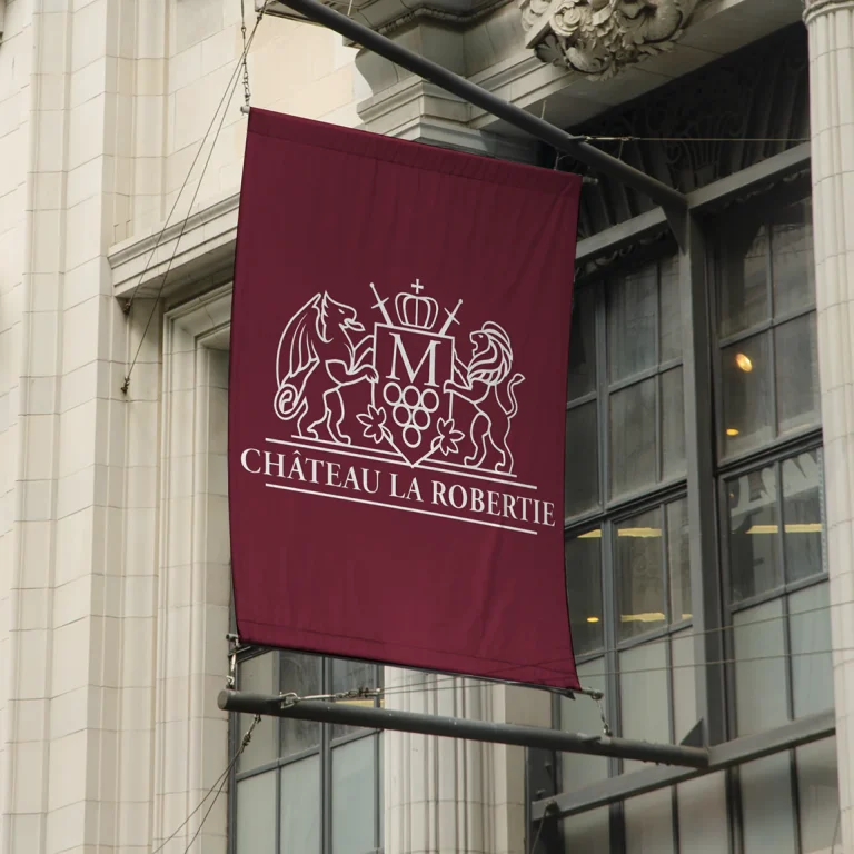 Висящо бордо знаме с бяло лого на Château La Robertie, монтирано на метална конструкция пред сграда с класическа архитектура.